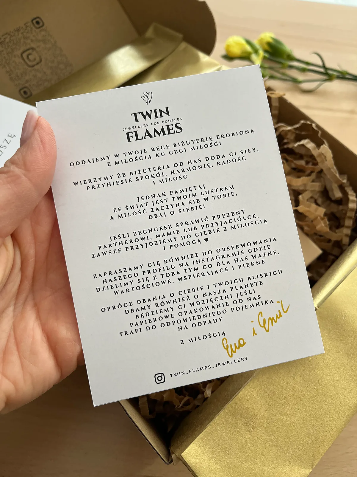 Dłoń trzyma kartę podziękowania Twin Flames nad otwartym pudełkiem ze złotym papierem i żółtymi kwiatami